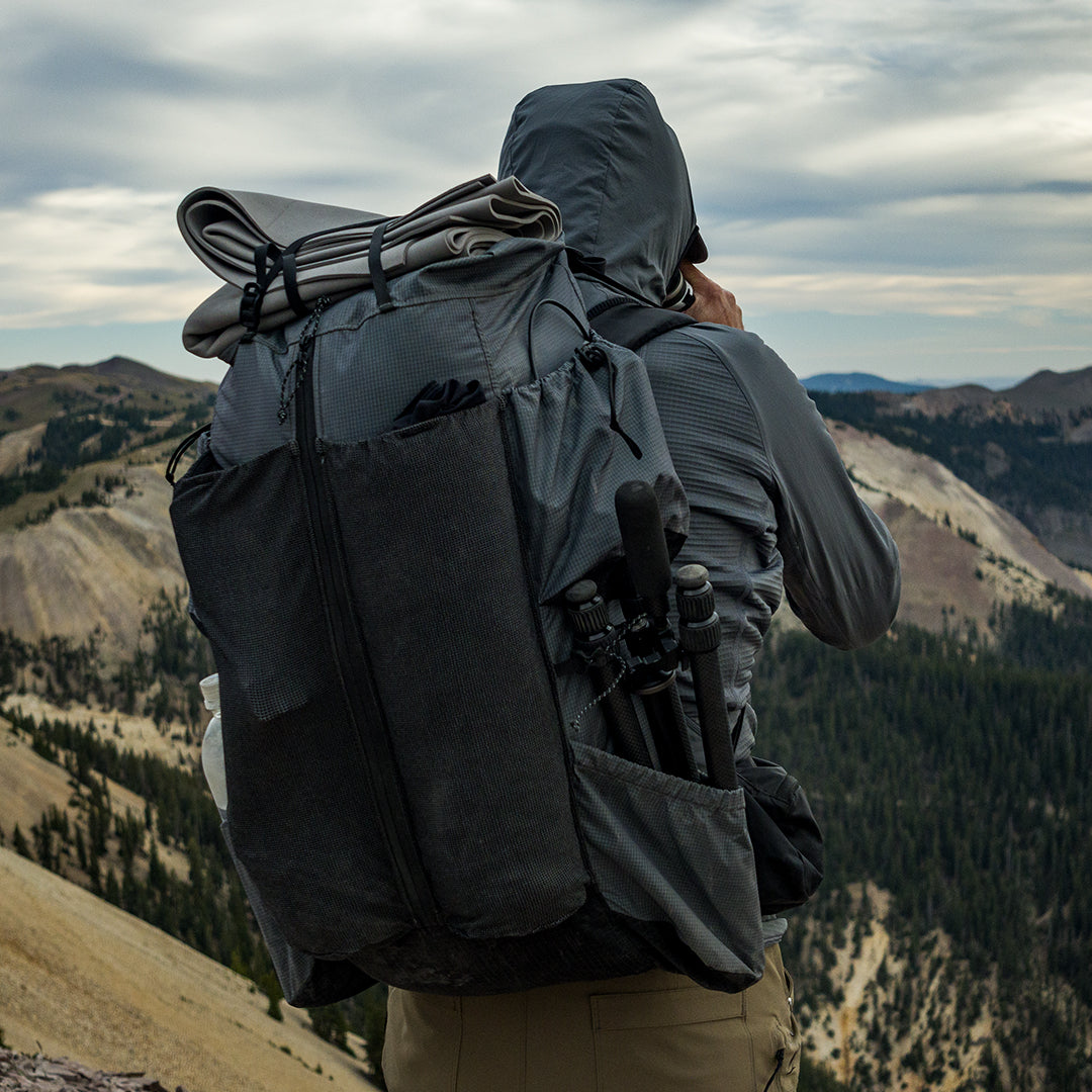 Hiker wearing a Outdoor Vitals Shadowlight backpack with gear strapped to the top and tripod stored in the side pocket, overlooking a rugged mountain ridgeline under cloudy skies.