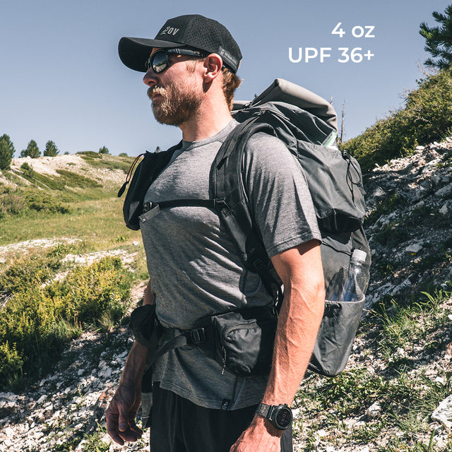 A man hiking outdoors wearing a wool t-shirt with a backpack, set against a rocky, grassy mountain landscape. White text in the upper right reads '4 oz UPF 36+'.