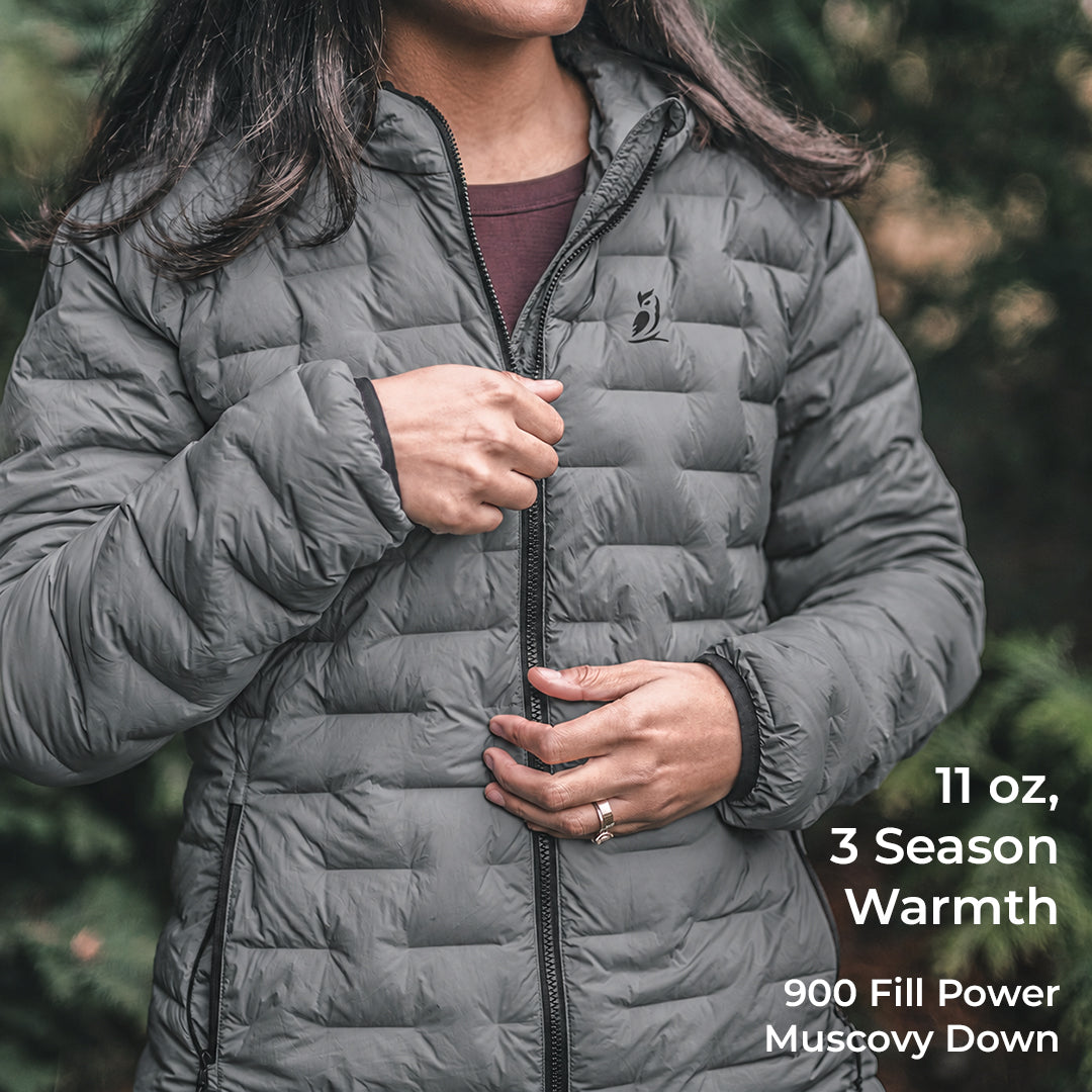 A woman wearing a dark gray quilted puffer jacket, shown from the chest down, with text overlays on the right detailing its features: '11 oz, 3 Season Warmth, 900 Fill Power Muscovy Down'.