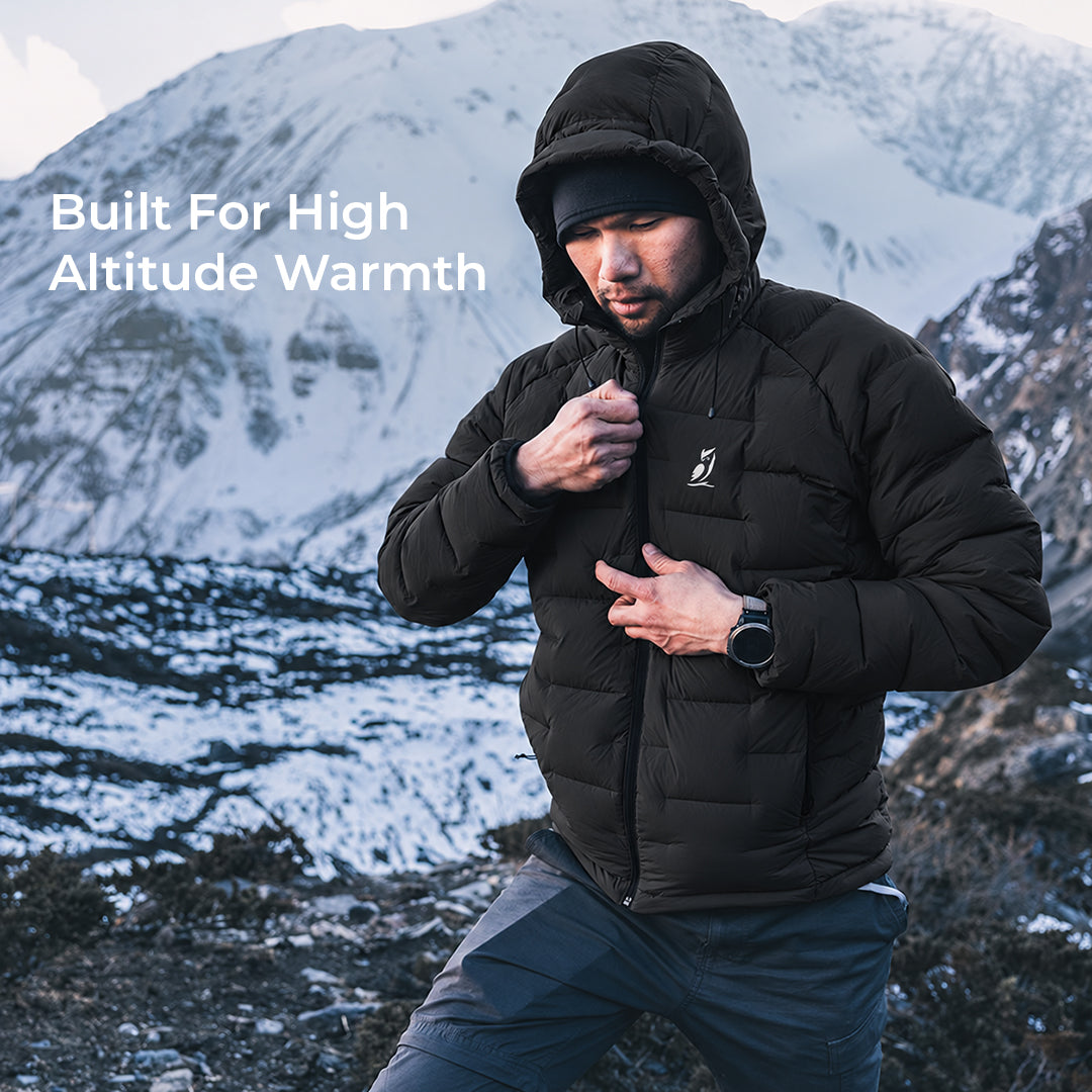 A man wearing a black hooded puffer jacket stands in a snowy mountain landscape, with the text 'Built For High Altitude Warmth' on the left side of the image.