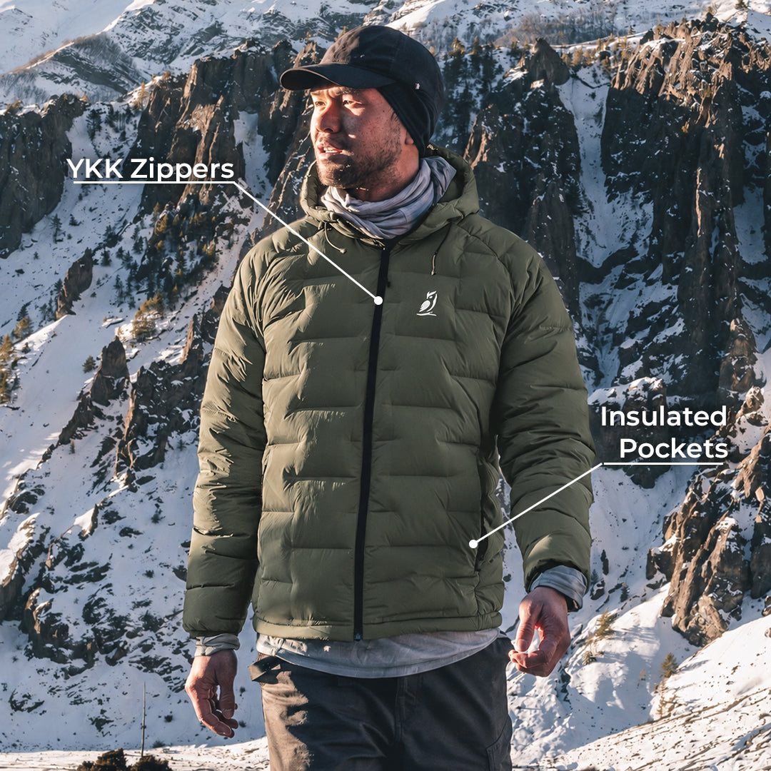A man wearing a green insulated puffer jacket stands in a snowy mountain landscape. White text labels point to the jacket, reading 'YKK Zippers' near the chest zipper area and 'Insulated Pockets' near the lower pocket area.